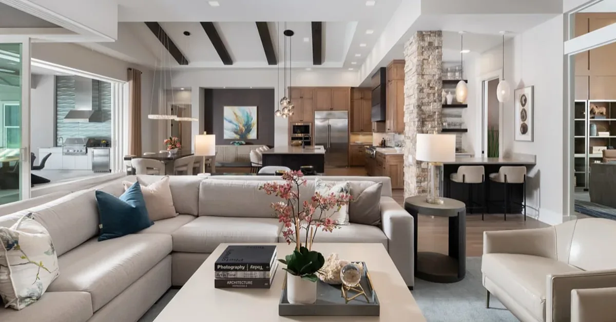 Modern open-concept living space features neutral sectional sofa, exposed ceiling beams, and seamless kitchen integration with natural stone accents.