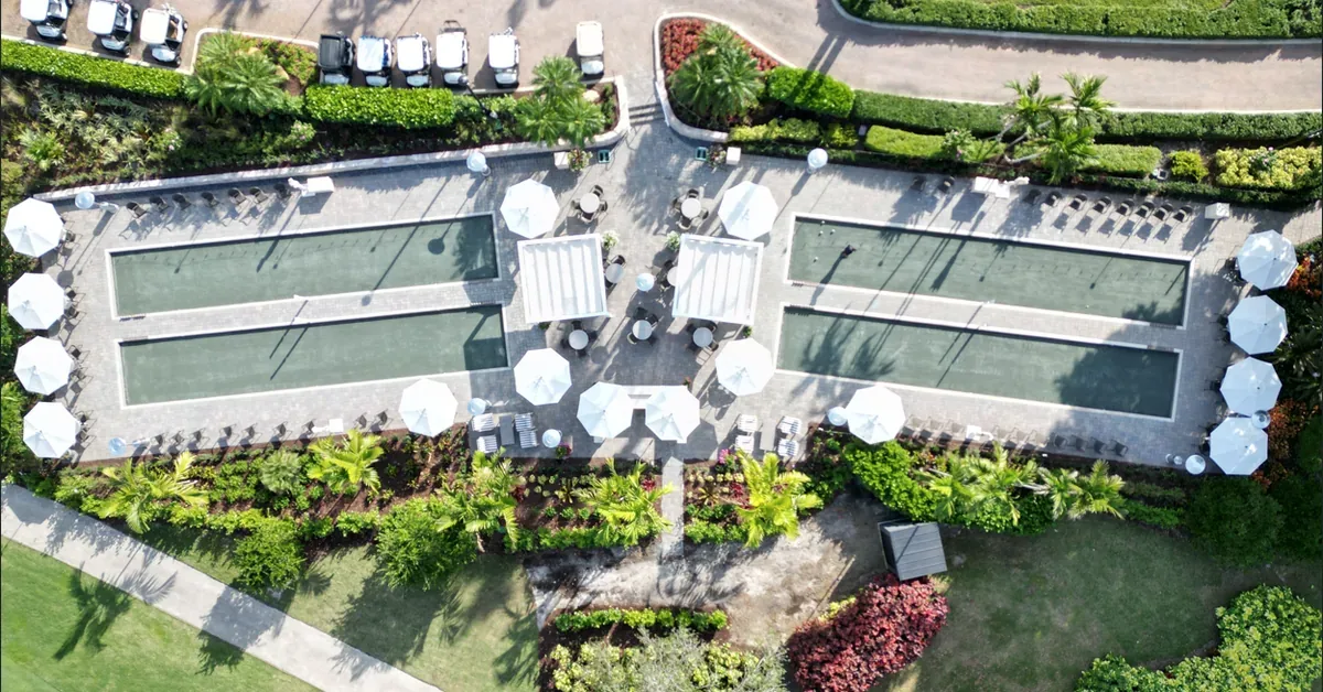 Aerial view of four manicured bocce ball courts with white market umbrellas, lush tropical landscaping, and golf cart parking at an upscale resort community.