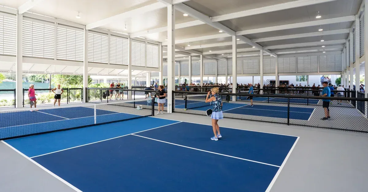 Covered indoor pickleball courts with blue playing surfaces and white lines, featuring multiple players enjoying games in a modern, open-air facility.
