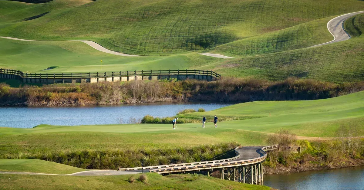Golfers play on a scenic course featuring rolling fairways, a tranquil lake, and wooden bridges amid lush green landscaping.