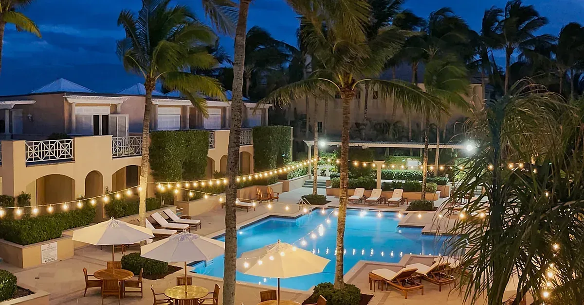 Luxury resort-style pool area glowing with string lights at dusk, surrounded by palm trees, lounge chairs, and elegant Caribbean-style villas.