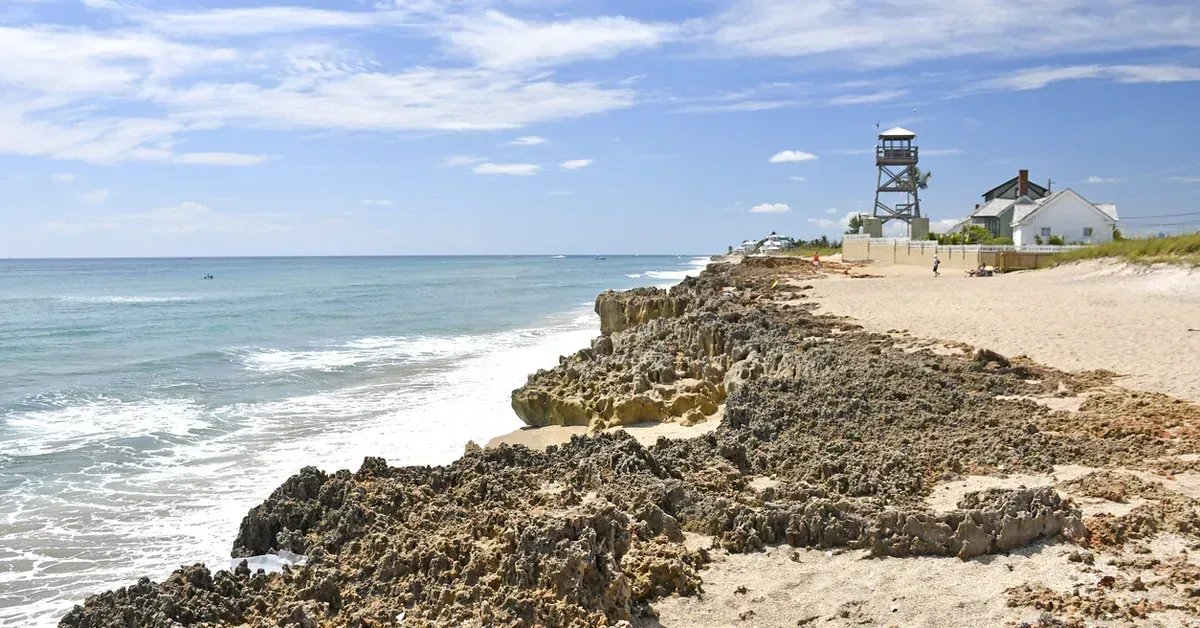 Rugged coquina rock formations line the shoreline at a scenic Florida beach community, with a lifeguard tower and coastal homes visible in the background.