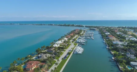 Aerial view of a luxury waterfront community featuring a private marina, elegant homes with terracotta roofs, palm trees, and stunning turquoise waters.